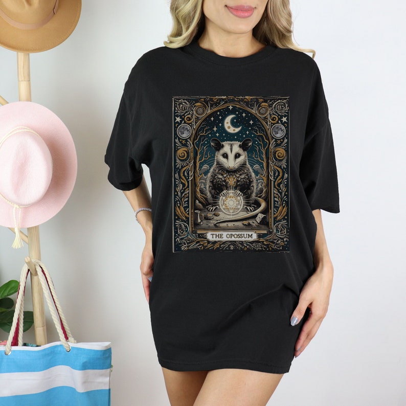 May include: Black t-shirt with a graphic print of an opossum holding a crystal ball. The graphic is surrounded by a gold frame with the text "THE OPOSSUM".