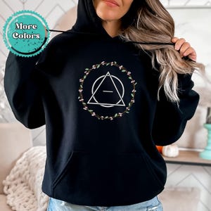 May include: Black hooded sweatshirt with a white geometric design inside a wreath of green leaves and pink flowers. The design features a circle with a triangle inside and a horizontal line across the center.