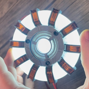 May include: A glowing, circular prop with a central light source, surrounded by a gray, segmented frame and copper-colored coils. The prop is held in a hand, with red wires visible, suggesting a power source. The design is reminiscent of a fictional energy device.