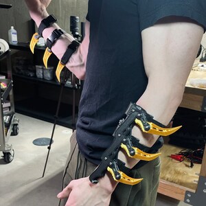 Puede incluir: Un brazalete negro para el brazo con tres garras de metal amarillo unidas. El brazalete se lleva en el brazo izquierdo y tiene una correa negra.