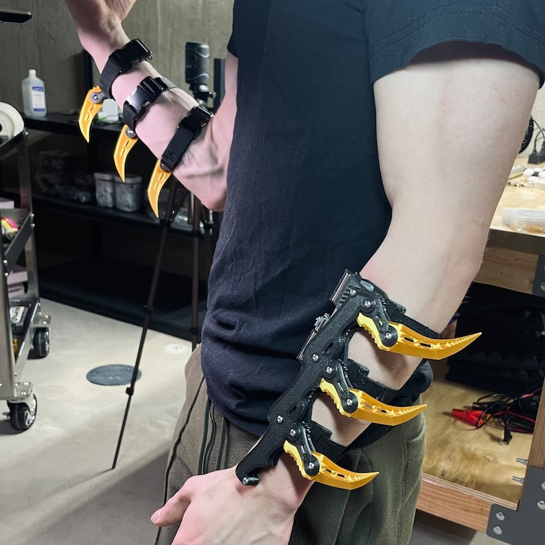 Puede incluir: Un brazalete negro para el brazo con tres garras de metal amarillo unidas. El brazalete se lleva en el brazo izquierdo y tiene una correa negra.