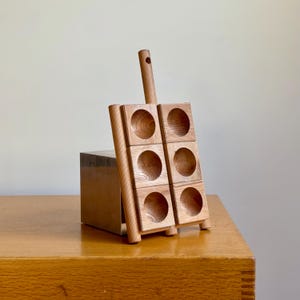 May include: A wooden wine bottle holder with six circular openings. The holder is made of light-colored wood and has a handle at the top. It is leaning against a wooden box on a wooden table.