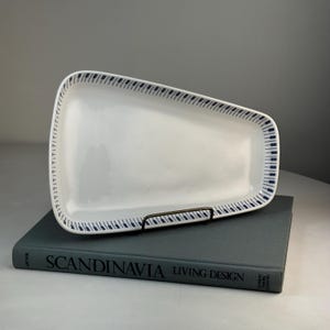 May include: A rectangular white ceramic serving platter with a blue patterned border. The platter is displayed on a dark metal stand, resting on a book titled "SCANDINAVIA LIVING DESIGN". The design features a series of blue, stylized shapes.