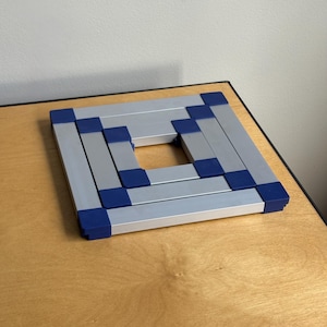 May include: A square, expandable trivet constructed from silver-colored metal bars connected by dark blue plastic corner joints. The frame features a hollow center and a geometric, modular design with a flat, low-profile structure.