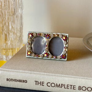 May include: A rectangular double photo frame with a metallic finish, featuring two oval openings. The border is decorated with a series of small, clear rhinestones and larger, iridescent red-toned circular crystals arranged in a repeating pattern.
