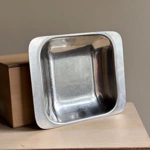 May include: A square, silver-colored metal bowl with a wide, flat rim and rounded corners. The interior surface of the bowl is reflective and shows light scratches, while the exterior rim has a smooth, matte metallic finish.