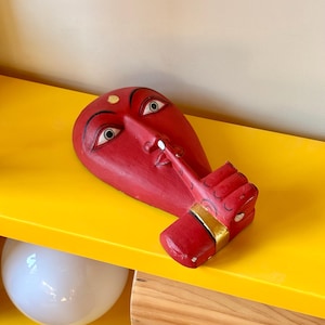 May include: A red wooden mask with a face design, featuring white eyes, a gold dot on the forehead, and a finger touching the nose. The mask is resting on a yellow shelf, with a white sphere and wooden block visible below.