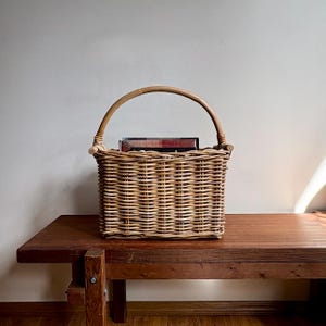 May include: A rectangular woven wicker basket with a curved handle, resting on a dark wooden bench. The basket is filled with books. The basket and handle are light brown, and the bench is a rich, dark brown. The background is a plain, off-white wall.