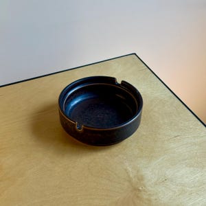 May include: A round, dark brown ashtray with a glossy finish. The ashtray has a small lip for resting cigarettes and a contrasting gold-colored rim. It sits on a light-colored wooden surface.