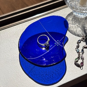 May include: A small, irregular, cobalt blue glass trinket dish featuring a thin white line design and a central silver-toned ring with a clear stone. The dish has a smooth, glossy finish and casts a deep blue shadow on the surface below it.