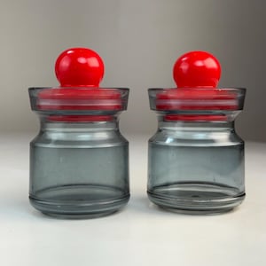 May include: Two matching glass jars with gray bodies and red lids. Each jar has a round red knob on top of the lid. The jars are cylindrical with a slightly wider base and a narrower neck.