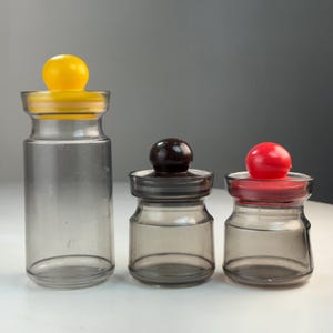 May include: Three glass jars with colored lids and spherical knobs. The jars vary in size, with a yellow lid on the tallest jar, a brown lid on the middle jar, and a red lid on the smallest jar. The jars are clear with a gray tint.