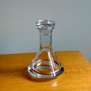 May include: A clear glass vase with a wide, rounded base and a narrow neck. The vase has a flared shape and a thick, rounded rim. It sits on a wooden surface against a neutral gray background. The vase is empty and reflects light.