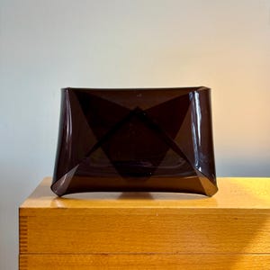 May include: A dark brown, geometric-shaped decorative object. The object has a rectangular base with angled sides and a diamond-shaped design in the center. The object is made of a translucent material and sits on a wooden surface.