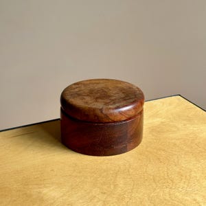 May include: A round, wooden box with a lid. The box is made of dark brown wood with a smooth, polished finish. The lid sits flush on top of the box. The box is sitting on a light-colored wooden surface.