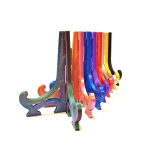 May include: A row of colorful display stands in various hues, including iridescent, green, orange, pink, blue, purple, yellow, and black. Each stand has a curved design with a base and a support for displaying items. The stands are arranged against a white background.
