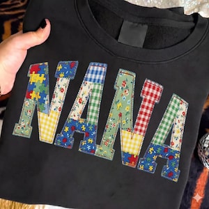 May include: Black sweatshirt with the word "NANA" in colorful patchwork letters. The letters feature various patterns, including puzzle pieces, floral prints, and gingham, creating a vibrant and personalized design.