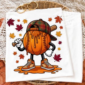 Op de afbeelding: Een wit t-shirt met een afbeelding van een pompoen-honkbalpersonage dat een geruite honkbalpet en hoge sneakers draagt en een houten honkbalknuppel vasthoudt. Het personage is omringd door verspreide rode, oranje en gele herfstbladeren.