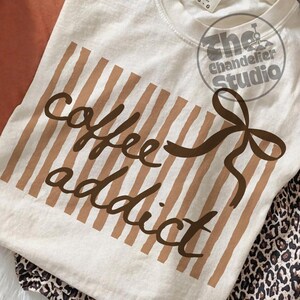 May include: Cream-colored sweatshirt with the words "coffee addict" in brown script. The design includes brown vertical stripes and a brown bow. The Chandelier Studio logo is in the upper right corner. The shirt is folded on a surface with a leopard print.