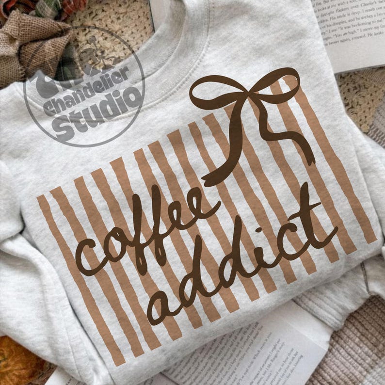 May include: A light gray sweatshirt with a brown bow and vertical striped design. The words "coffee addict" are written in a brown script font across the front. The sweatshirt is displayed on a neutral background.