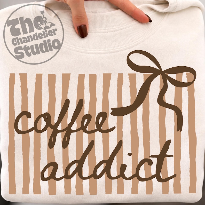 May include: Cream-colored sweatshirt with the text "coffee addict" in a brown script font. The design includes vertical brown stripes and a brown bow. The Chandelier Studio logo is in the upper left corner.