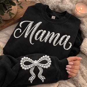 Puede incluir: Sudadera negra con la palabra "Mama" escrita con perlas blancas. Un lazo de perlas está en la manga. La sudadera está sobre una superficie suave y texturizada.