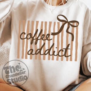 May include: Cream-colored sweatshirt with the text "coffee addict" in brown script. The design includes vertical brown stripes and a brown bow. The Chandelier Studio logo is at the bottom.