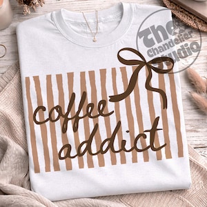 May include: White t-shirt with brown vertical stripes and the words "coffee addict" in a brown script font. A brown bow is on the upper right. The Chandelier Studio logo is in the upper right corner.