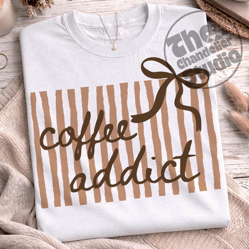 May include: White t-shirt with brown vertical stripes and the words "coffee addict" in a brown script font. A brown bow is on the upper right. The Chandelier Studio logo is in the upper right corner.