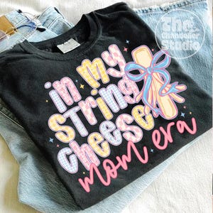 May include: Black t-shirt with the text "in my string cheese mom era" in pastel colors. The design includes a string cheese illustration with a blue bow. The shirt is folded on top of a pair of blue jeans.