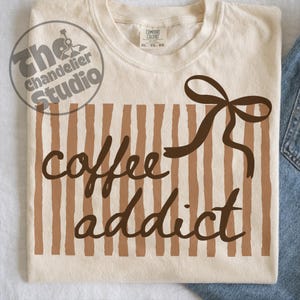 May include: Cream-colored t-shirt with a brown vertical striped pattern and the words "coffee addict" in a brown script font. A brown bow is at the top right. The shirt is a Comfort Colors XL.