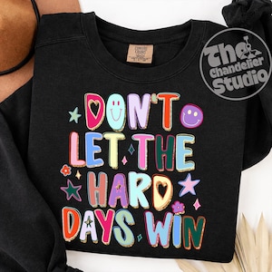 May include: Black sweatshirt with the text "DON'T LET THE HARD DAYS WIN" in colorful, outlined letters. The design includes stars, hearts, and a smiley face. The sweatshirt has a Comfort Colors tag.