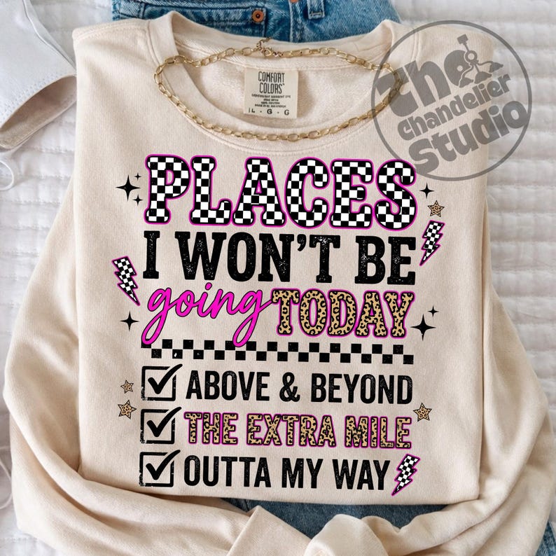 May include: Cream-colored sweatshirt with the text "PLACES I WON'T BE going TODAY" in black, pink, and leopard print. The design includes lightning bolts, stars, and checkmarks with phrases like "ABOVE & BEYOND". A gold chain necklace is draped on top.