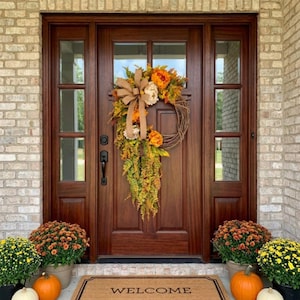 May include: A brown wooden front door with a wreath featuring orange and yellow flowers, green foliage, and a tan burlap bow. A tan welcome mat sits on the floor, flanked by two potted orange chrysanthemum plants and two potted yellow chrysanthemum plants.