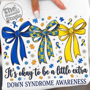 May include: White sweatshirt with a design featuring three large bows in blue, yellow, and blue/yellow puzzle piece patterns. The text "It's okay to be a little extra" and "DOWN SYNDROME AWARENESS" are also printed on the shirt.