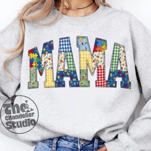Peut inclure: Un sweat-shirt gris clair avec le mot "MAMA" en grandes lettres de style patchwork. Les lettres sont remplies de divers motifs, notamment des motifs floraux, vichy et en forme de pièces de puzzle. Le logo The Chandelier Studio est visible en bas à gauche.
