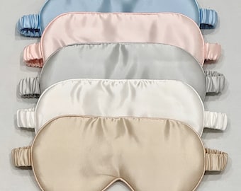 May include: A stack of six silk sleep masks in various pastel colors: light blue, pink, gray, white, and beige. Each mask has an elastic band for a comfortable fit. The masks are arranged in a neat, overlapping row.