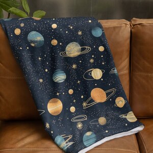 May include: A soft, plush throw blanket with a navy blue background and a repeating pattern of gold and blue planets and stars.