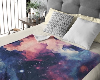 Puede incluir: Una manta suave y mullida con estampado de galaxia que presenta nubes giratorias de color rosa, azul y morado con estrellas blancas. La manta está sobre una cama con sábanas blancas y almohadas grises.