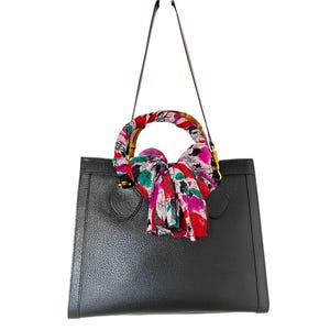 May include: Black leather tote bag with a bamboo handle and a colorful floral scarf tied around it. The bag has a rectangular shape and a long, thin shoulder strap. The scarf features red, pink, green, and white floral patterns.
