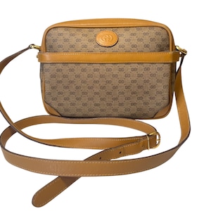 May include: A tan and brown crossbody bag with a repeating pattern. The bag has a tan leather strap and trim, and a gold-colored buckle. A circular emblem is centered on the front of the bag.