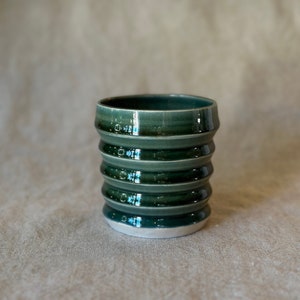 Könnte beinhalten: Eine grüne Keramiktasse mit einem strukturierten, gerippten Design. Die Tasse hat einen weißen Rand und eine glänzende Oberfläche.
