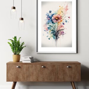 May include: A watercolor painting of a colorful flower bouquet with pink, blue, yellow, and green hues. The flowers are arranged in a loose, organic style, and the background is a soft, white wash. The painting is framed in a white frame and is hanging on a white wall.