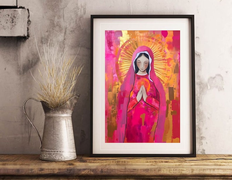 May include: A framed painting of a woman in a pink robe with a golden halo. The woman is praying with her hands clasped together. The background is a mix of pink, gold, and white.
