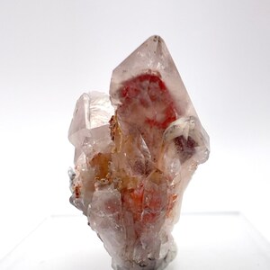 May include: A cluster of clear quartz crystals with red inclusions. The crystals are clustered together in a natural formation.