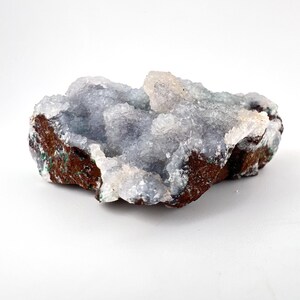 Puede incluir: Un esp&eacute;cimen de geoda natural con c&uacute;mulos de cristales azul claro. La formaci&oacute;n de cristales se asienta sobre una base marr&oacute;n oscuro y rugosa con toques verdes. La geoda se muestra sobre un fondo blanco.