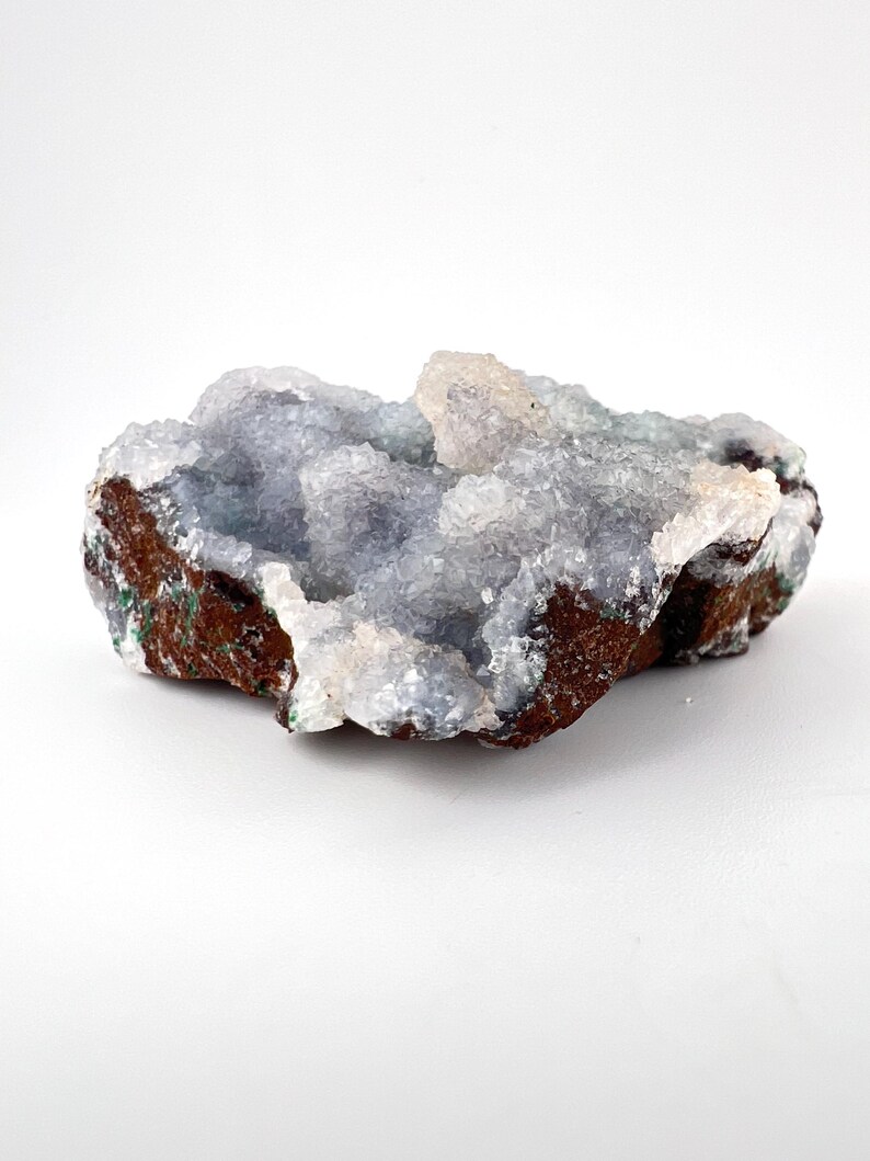 Puede incluir: Un esp&eacute;cimen de geoda natural con c&uacute;mulos de cristales azul claro. La formaci&oacute;n de cristales se asienta sobre una base marr&oacute;n oscuro y rugosa con toques verdes. La geoda se muestra sobre un fondo blanco.