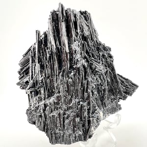 May include: A striking black stibnite mineral specimen. The crystal formation features numerous, elongated, needle-like structures radiating from a central point. The mineral is displayed on a clear acrylic stand against a white background.