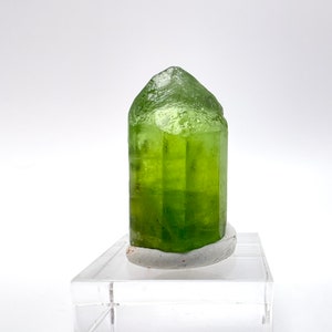 May include: A green crystal point on a clear acrylic display stand. The crystal is a deep green color and has a pointed tip. The crystal is about 3 inches tall.