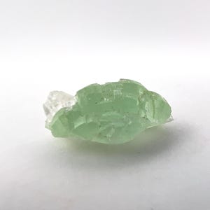 May include: A raw, light green fluorite crystal with a translucent appearance. The crystal has a rough, uneven texture and a small, clear quartz formation attached to one side. The fluorite is a pale, minty green color.